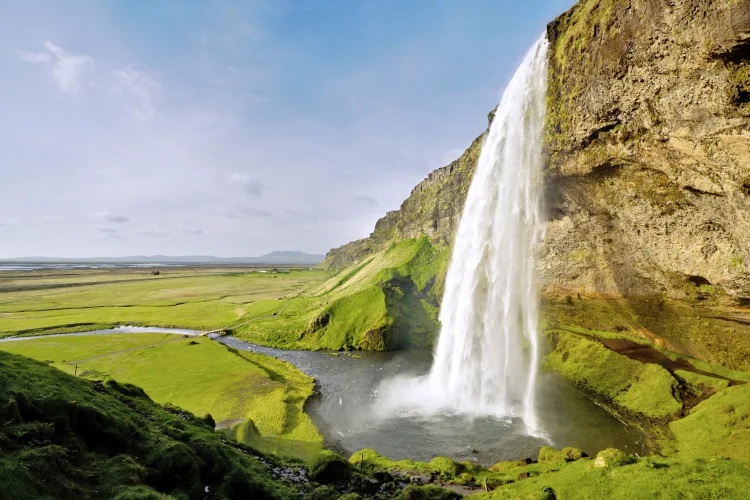 Ein hoher, schmaler Wasserfall stürzt von einer steilen, grasbewachsenen Klippe in ein Becken, umgeben von weiten grünen Feldern unter blauem Himmel.