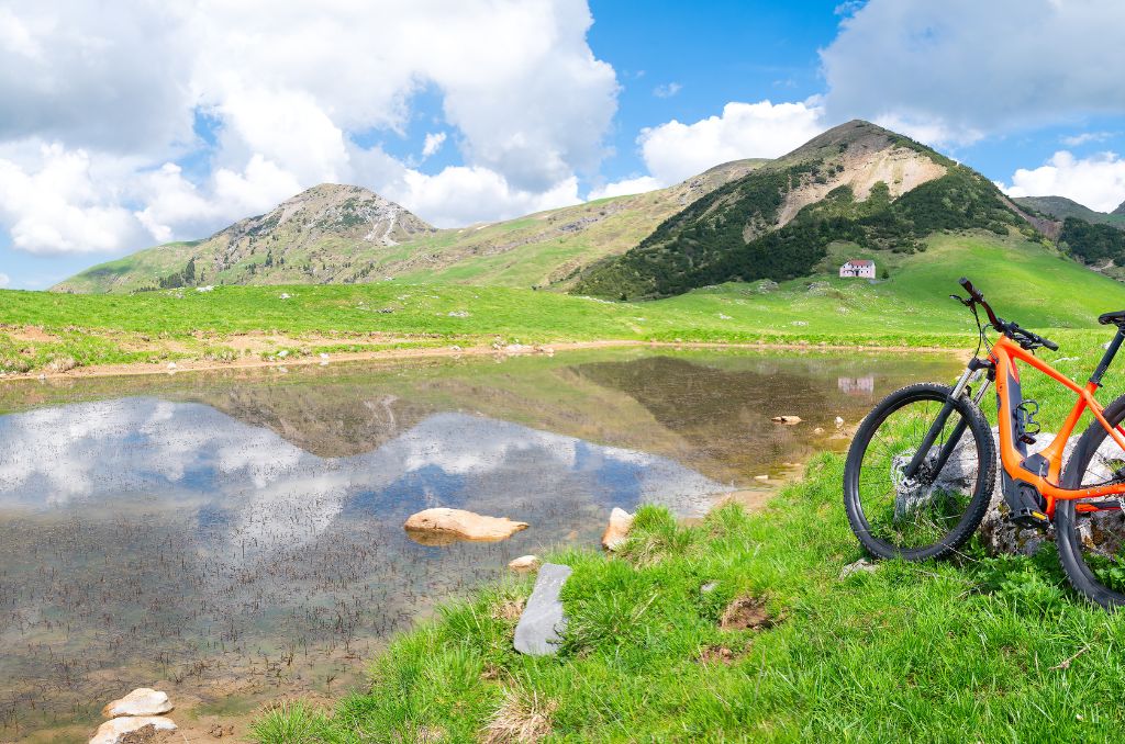 An einem Bergsee-Ufer steht ein orangefarbenes E-Mountainbike. Die Berge und der Wolkenhimmel spiegeln sich im Wasser.